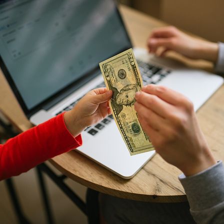 Child hands dollar bill to adult at a table with a laptop and paperwork in a home setting