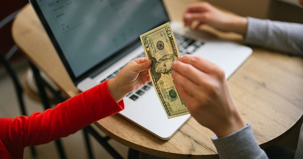 Child hands dollar bill to adult at a table with a laptop and paperwork in a home setting