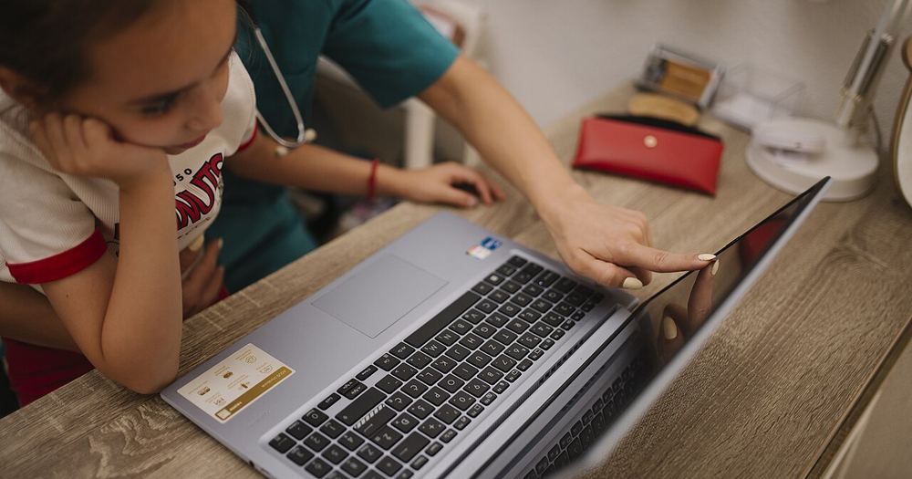 Child interacts with healthcare professional while exploring medical software on a laptop