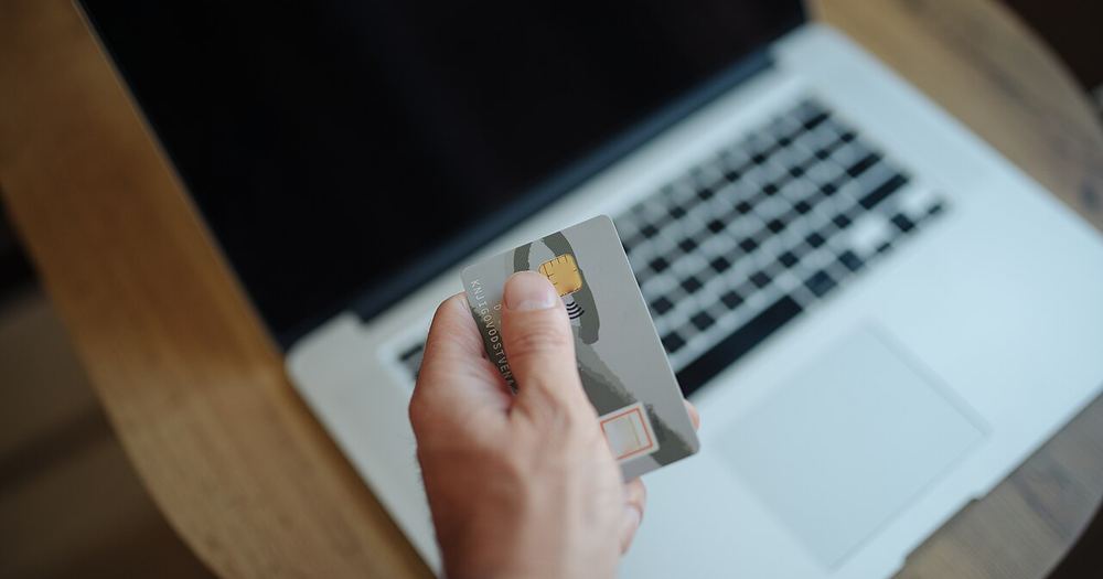 Person holds a credit card while using a laptop on a wooden table in a home or office