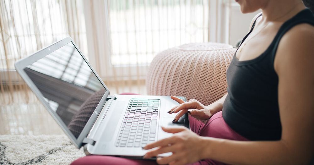 A woman in a black tank top sits on a soft floor, focusing on her laptop